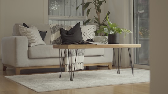 A cozy living room interior featuring a light-colored sofa adorned with various patterned cushions. A wooden coffee table with metal legs is centered on a soft, neutral-colored rug. A potted plant and a blanket add a touch of greenery and warmth. The room is well-lit with natural light streaming through a window.