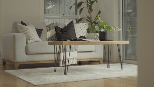 A cozy living room interior featuring a light-colored sofa adorned with various patterned cushions. A wooden coffee table with metal legs is centered on a soft, neutral-colored rug. A potted plant and a blanket add a touch of greenery and warmth. The room is well-lit with natural light streaming through a window.