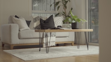 A cozy living room interior featuring a light-colored sofa adorned with various patterned cushions. A wooden coffee table with metal legs is centered on a soft, neutral-colored rug. A potted plant and a blanket add a touch of greenery and warmth. The room is well-lit with natural light streaming through a window.