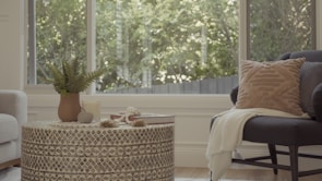 A cozy and stylish living room with a round patterned coffee table. On the table, there's a plant in a brown pot, a candle, a stone, and a stack of books. A soft armchair with a textured cushion and blanket is placed nearby, with large windows in the background allowing natural light to spill in.