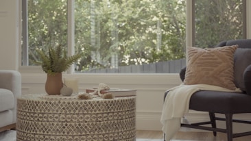 A cozy and stylish living room with a round patterned coffee table. On the table, there's a plant in a brown pot, a candle, a stone, and a stack of books. A soft armchair with a textured cushion and blanket is placed nearby, with large windows in the background allowing natural light to spill in.