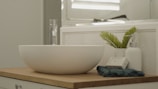 A minimalist bathroom countertop featuring a white vessel sink with a sleek, chrome faucet. Beside the sink, there is a potted plant with green leaves, a white textured soap dispenser, and a bar of natural soap wrapped in paper. The surface is wooden, and the background includes a decorative tile wall and a mirror reflecting light.
