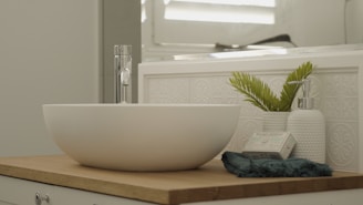 A minimalist bathroom countertop featuring a white vessel sink with a sleek, chrome faucet. Beside the sink, there is a potted plant with green leaves, a white textured soap dispenser, and a bar of natural soap wrapped in paper. The surface is wooden, and the background includes a decorative tile wall and a mirror reflecting light.
