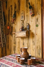 A rustic interior scene featuring a wooden wall adorned with decorative items. Two wine bottles in fabric holders hang on the wall alongside small wooden crafts and a small statue. A woven basket containing an orange fruit is mounted on the left side. A wooden log shelf displays a small ceramic pot and grape decoration. In the foreground, a clay pitcher and matching cups are arranged on a patterned textile tablecloth.