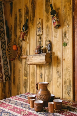 A rustic interior scene featuring a wooden wall adorned with decorative items. Two wine bottles in fabric holders hang on the wall alongside small wooden crafts and a small statue. A woven basket containing an orange fruit is mounted on the left side. A wooden log shelf displays a small ceramic pot and grape decoration. In the foreground, a clay pitcher and matching cups are arranged on a patterned textile tablecloth.