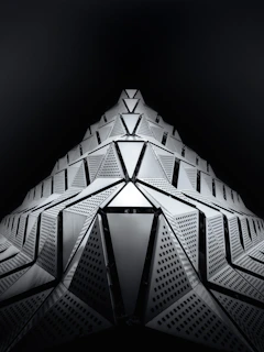 A futuristic architectural structure with geometric shapes extending upward in a symmetrical pattern. The surface is composed of triangular and rectangular panels with intricate perforations, creating a dynamic and sleek appearance against a dark background.