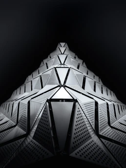 A futuristic architectural structure with geometric shapes extending upward in a symmetrical pattern. The surface is composed of triangular and rectangular panels with intricate perforations, creating a dynamic and sleek appearance against a dark background.