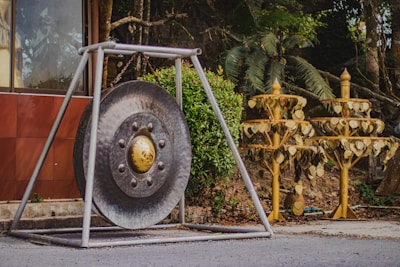 A large metal gong is mounted on a triangular stand situated outdoors. Nearby, decorative golden tree structures adorned with heart-shaped leaves stand on the right. The scene includes a lush backdrop of greenery and part of a building wall on the left.