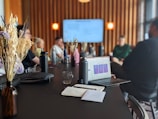 A group of people sitting around a long black table, engaged in a meeting or presentation. Several personal items like water bottles and notebooks are visible on the table. A laptop with an open presentation is prominently placed in the foreground. The room features modern decor with wooden wall panels and soft lighting from visible lamps. A large screen at the end of the table displays a presentation slide.