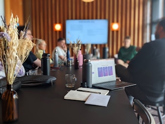 A group of people sitting around a long black table, engaged in a meeting or presentation. Several personal items like water bottles and notebooks are visible on the table. A laptop with an open presentation is prominently placed in the foreground. The room features modern decor with wooden wall panels and soft lighting from visible lamps. A large screen at the end of the table displays a presentation slide.