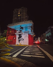 The image captures a vibrant urban scene at night, featuring a prominent building with bold street art. The building is painted in red and white, with a large mural depicting a person on its facade. A towering structure is visible in the background, illuminated by scattered lights. The foreground shows a street with painted signage and vegetation on the side.