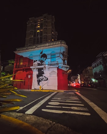 The image captures a vibrant urban scene at night, featuring a prominent building with bold street art. The building is painted in red and white, with a large mural depicting a person on its facade. A towering structure is visible in the background, illuminated by scattered lights. The foreground shows a street with painted signage and vegetation on the side.