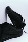 A pair of black sneakers with a sleek design is positioned against a white background. One sneaker is slightly elevated and leaning against a surface, while the other lies flat with laces partially untied.
