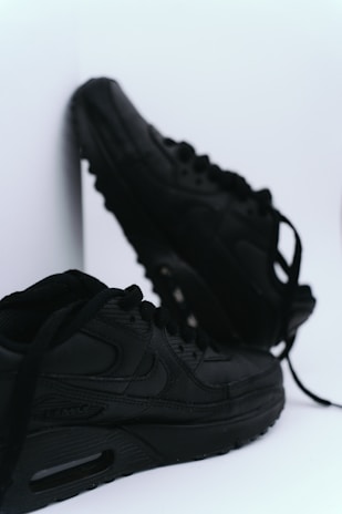 A pair of black sneakers with a sleek design is positioned against a white background. One sneaker is slightly elevated and leaning against a surface, while the other lies flat with laces partially untied.