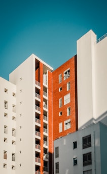 Tall, modern apartment buildings with a combination of white and red brick facades against a clear blue sky. The architectural design features balconies and windows with variations in patterns and textures.