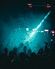 A crowd of people is gathered at a lively event under a dim, blue-green light. Confetti fills the air while a spotlight shines down from above, creating an energetic atmosphere. Silhouettes of hands and heads can be seen among the crowd, indicating that the participants are actively enjoying the moment.