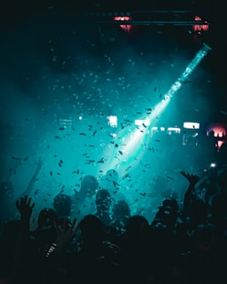 A crowd of people is gathered at a lively event under a dim, blue-green light. Confetti fills the air while a spotlight shines down from above, creating an energetic atmosphere. Silhouettes of hands and heads can be seen among the crowd, indicating that the participants are actively enjoying the moment.
