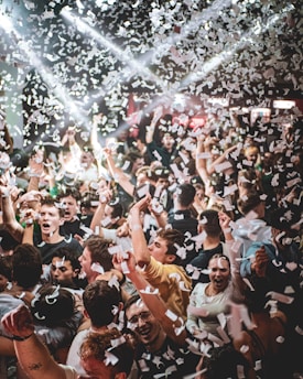 A crowded party scene with people dancing and throwing white confetti into the air. Intense lighting, including bright spotlights, adds energy to the festive atmosphere.