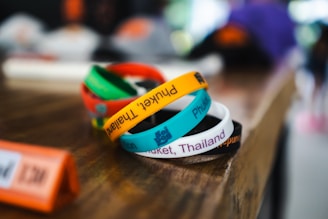 Colorful wristbands are stacked on a wooden table. They have text printed on them, with phrases like 'Phuket, Thailand'. The background is blurred, creating a shallow depth of field effect.