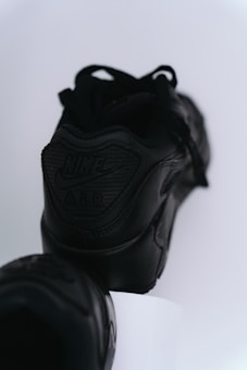 A pair of black Nike sneakers with a prominent logo on the heel against a minimalistic white background. The focus is on the embossed 'Nike Air' branding, highlighting the sleek and modern design of the shoes.