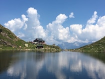 A serene mountain landscape with a calm lake reflecting the sky and surrounding greenery. A lodge sits on the grassy bank surrounded by small hills, and fluffy white clouds float in the blue sky above distant mountains.