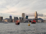 Several traditional Chinese junks with bright red sails are sailing on a calm water body, with a modern cityscape featuring tall skyscrapers in the background. The sky is partly cloudy, and there is a buoy visible in the water.