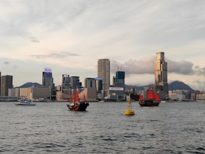 Several traditional Chinese junks with bright red sails are sailing on a calm water body, with a modern cityscape featuring tall skyscrapers in the background. The sky is partly cloudy, and there is a buoy visible in the water.
