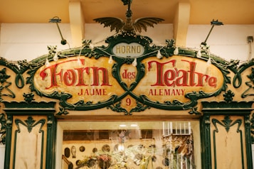 An ornate and decorative facade of a bakery or shop, featuring intricate designs and the words 'Horno des Teatre' prominently displayed in red and ornate script. The sign is bordered with elaborate green flourishes and decorative lighting. The stone wall and entrance add an antique and charming aesthetic to the scene.