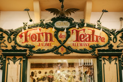 An ornate and decorative facade of a bakery or shop, featuring intricate designs and the words 'Horno des Teatre' prominently displayed in red and ornate script. The sign is bordered with elaborate green flourishes and decorative lighting. The stone wall and entrance add an antique and charming aesthetic to the scene.