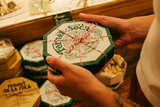 A person holds an octagonal green box with a festive red and white string tied around it. The box has intricate designs and text on the lid. Similar boxes are stacked in the background, creating a warm and inviting display.