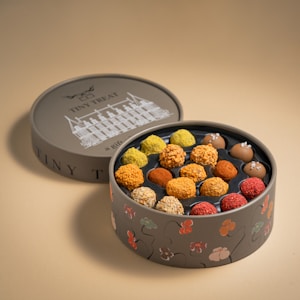A round, elegant box containing an assortment of colorful truffles. The box is decorated with a subtle floral pattern and a sketch of a building on the lid. The truffles vary in color, including shades of red, yellow, orange, green, and brown, and are arranged in a neat circular pattern.