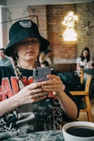 A person is sitting in a cafe, wearing a black bucket hat with a butterfly emblem and braids. They are focused on their smartphone. Behind them, there's a neon sign on a brick wall featuring a cartoonish character. Others are seated in the background, and a cup of coffee is in the foreground.