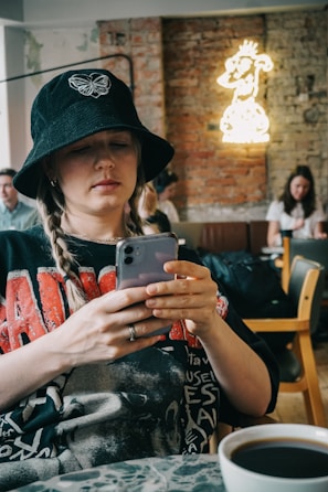 A person is sitting in a cafe, wearing a black bucket hat with a butterfly emblem and braids. They are focused on their smartphone. Behind them, there's a neon sign on a brick wall featuring a cartoonish character. Others are seated in the background, and a cup of coffee is in the foreground.