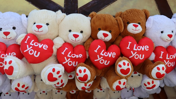 Several plush teddy bears are arranged in a row, each holding a red heart with 'I Love You' written on it. The bears are primarily in shades of white and brown, with red accents on their noses and paws, creating a cozy and affectionate display.