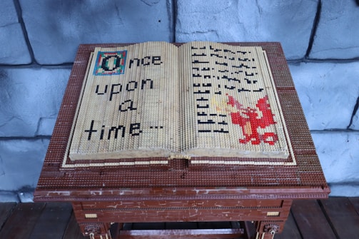 A large open book made entirely out of LEGO bricks sits on a wooden stand. The left page has the words 'Once upon a time...' crafted from black LEGO pieces. The right page features a red dragon made from LEGO bricks, with undecipherable text symbols in black surrounding it. The background consists of grey stone-like walls that resemble a medieval castle setting.