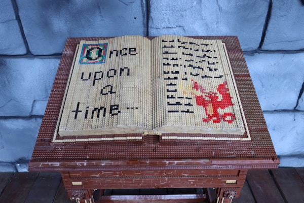 A large open book made entirely out of LEGO bricks sits on a wooden stand. The left page has the words 'Once upon a time...' crafted from black LEGO pieces. The right page features a red dragon made from LEGO bricks, with undecipherable text symbols in black surrounding it. The background consists of grey stone-like walls that resemble a medieval castle setting.