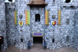 A stone castle facade with two large round towers and colorful banners featuring heraldic symbols. There are two shield emblems depicting a dragon and a lion on the towers. Two statues of knights in armor stand at the entrance. A small window with iron bars is visible, and grey stones form the bulk of the walls.