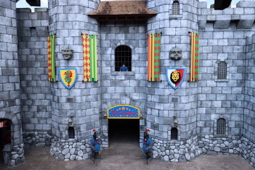 A stone castle facade with two large round towers and colorful banners featuring heraldic symbols. There are two shield emblems depicting a dragon and a lion on the towers. Two statues of knights in armor stand at the entrance. A small window with iron bars is visible, and grey stones form the bulk of the walls.