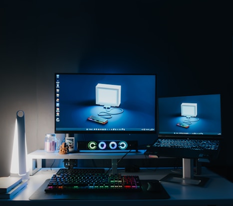 A modern computer workstation setup featuring two monitors, a colorful backlit mechanical keyboard, a sleek desk lamp, and various decorative items. The primary monitor has a retro-style image of a computer and keyboard with a blue background, while the secondary monitor displays the same image. The room is dimly lit, creating a focused and tech-centric atmosphere.