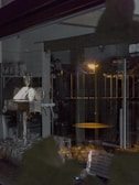 The image presents a dimly lit interior of a bar or café, with glassware neatly arranged on the counter. There is a metal beer tap with handles visible on the left side. The background includes a grid-tiled wall and shelves holding various supplies. A warm light from outside casts a faint glow through a transparent door or window, illuminating a round table.