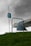A hospital bed with a weathered metal frame is seen placed outdoors on a grassy hill. An IV pole with medical equipment and IV bags stands beside the bed under an overcast sky. A blue blanket drapes over the edge of the bed, adding a touch of color to the muted scene.