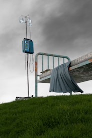 A hospital bed with a weathered metal frame is seen placed outdoors on a grassy hill. An IV pole with medical equipment and IV bags stands beside the bed under an overcast sky. A blue blanket drapes over the edge of the bed, adding a touch of color to the muted scene.