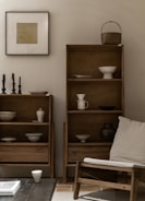 A minimalist living space featuring wooden shelves filled with ceramic bowls and vessels. A framed artwork is hung on the light grey wall. To the right, a wooden chair with a beige cushion is positioned nearby, along with a few candlesticks on a table. The aesthetic is calm and uncluttered.