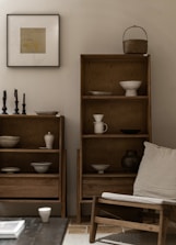 A minimalist living space featuring wooden shelves filled with ceramic bowls and vessels. A framed artwork is hung on the light grey wall. To the right, a wooden chair with a beige cushion is positioned nearby, along with a few candlesticks on a table. The aesthetic is calm and uncluttered.