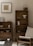 A minimalist living space featuring wooden shelves filled with ceramic bowls and vessels. A framed artwork is hung on the light grey wall. To the right, a wooden chair with a beige cushion is positioned nearby, along with a few candlesticks on a table. The aesthetic is calm and uncluttered.