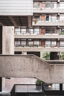 A concrete residential building with multiple stories, featuring balconies adorned with potted plants and flowers. The design is characterized by geometric lines and a brutalist architectural style. The shot captures the intersection of walkways and the texture of the concrete surfaces, creating a layered urban landscape.