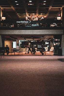 A dimly lit interior space features a martini bar on the upper level and an open area on the lower floor. People are sitting at tables and walking around in a modern, urban environment. The ceiling has a grid pattern with various colored panels.
