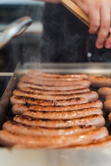 Several sausages are grilling on a hot plate with steam rising, while a hand using tongs is visible at the top.
