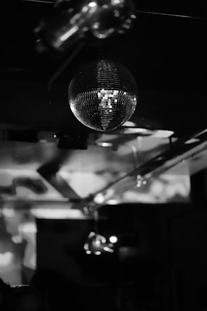 A black and white photograph featuring a disco ball suspended from the ceiling. The reflected light creates patterns on the walls, evoking a dynamic and lively ambiance. The room appears dimly lit, enhancing the contrast and texture of the disco ball and the surrounding shadows.