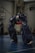 Two individuals wearing traditional kendo armor are engaged in a practice or match on an indoor court. They are holding long shinai swords, appearing to be in the midst of a sparring session. The setting is a sports hall with a basketball hoop visible in the background.